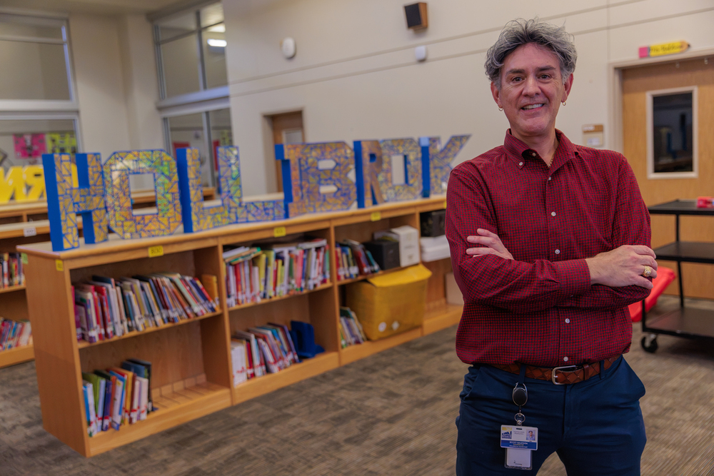 A man stands in a library with colorful "HOLLIBROOK" letters on the shelves behind him. He wears a red shirt and blue pants.