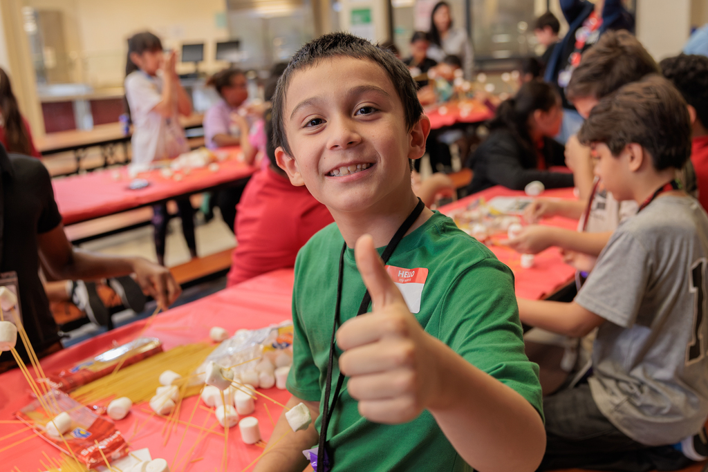 Kids sitting at tables, one boy wearing green gives two thumbs up. Tables covered with red cloth.