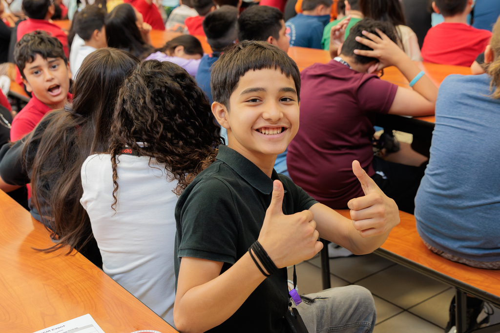 A young boy smiles and gives two thumbs up in a crowded room with other students.