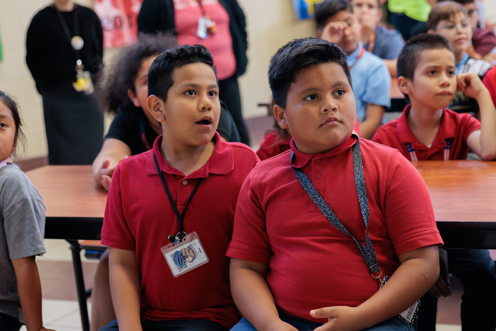 Two boys in red shirts seated at a table, looking forward. Behind them, children and an adult stand or sit.