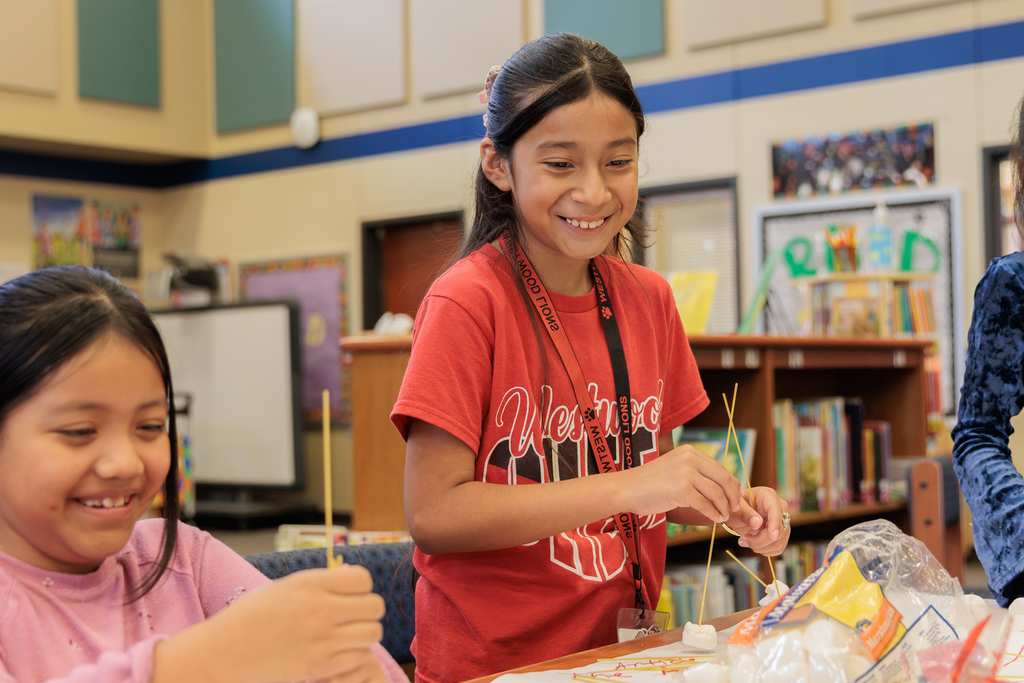 Two girls in a classroom, one holding sticks, the other smiling, with books on shelves and framed pictures in the background.