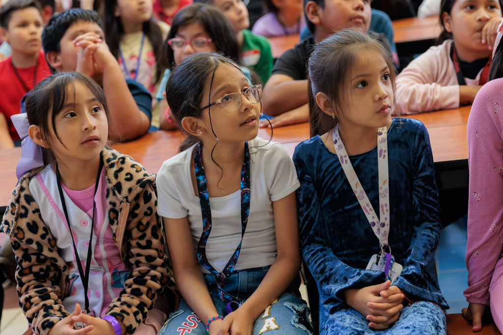 Several young girls wearing ID cards sit in a classroom. One girl in leopard print sits to the left.