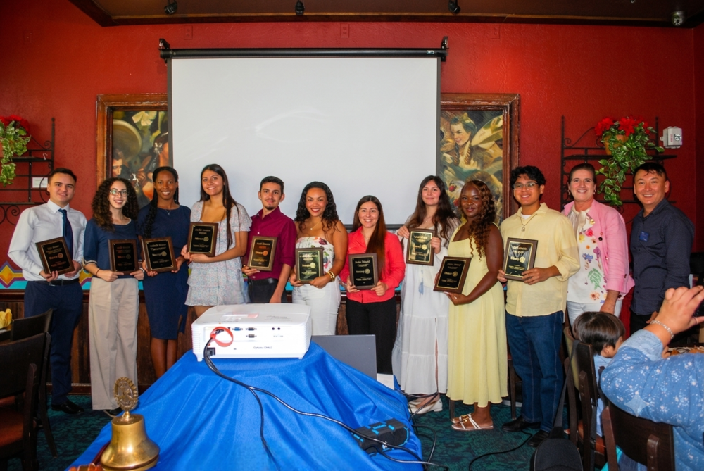 A group of individuals stands together, holding awards. They are in front of a red wall and a projector screen.