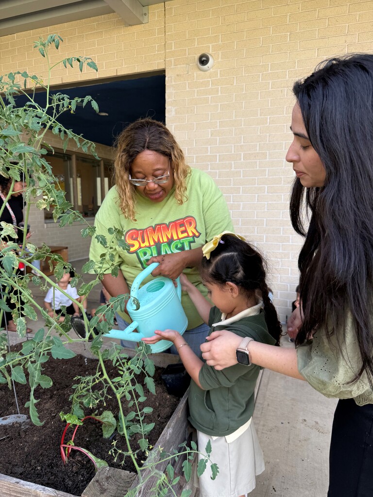 Two women and a young girl in a garden. The girl is watering a plant with a blue watering can.