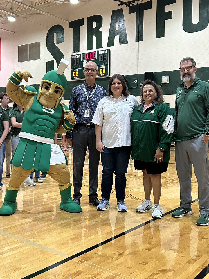 Four adults and a mascot stand on a basketball court in front of a wall.