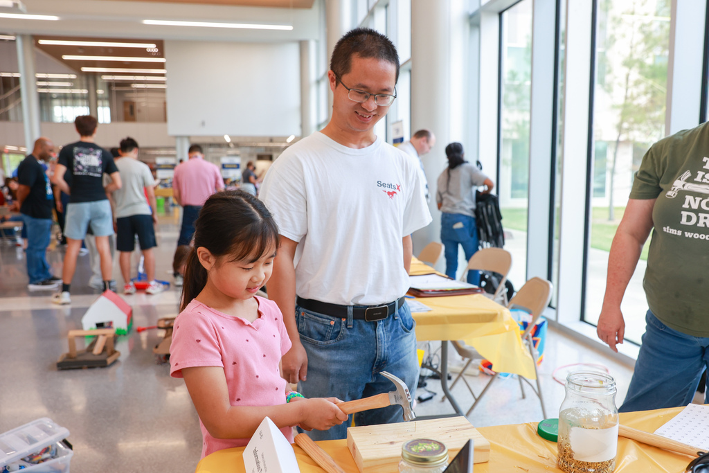 People in a room with tables, a girl uses a hammer on a wooden block, adults are nearby.