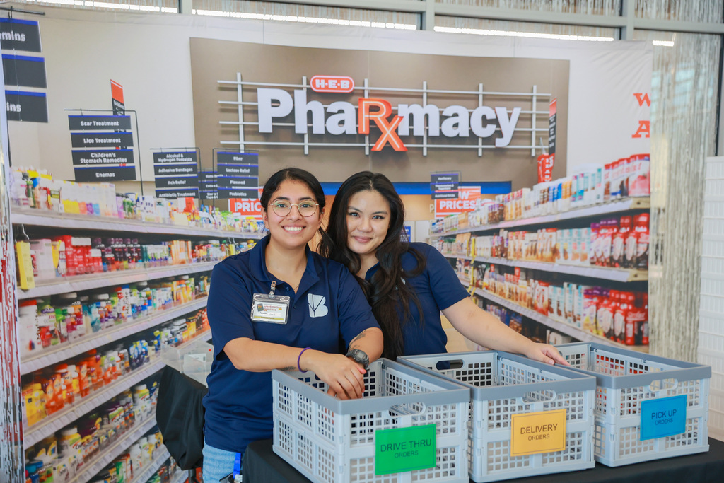 Two women in blue shirts stand behind a table with three storage bins in a pharmacy.