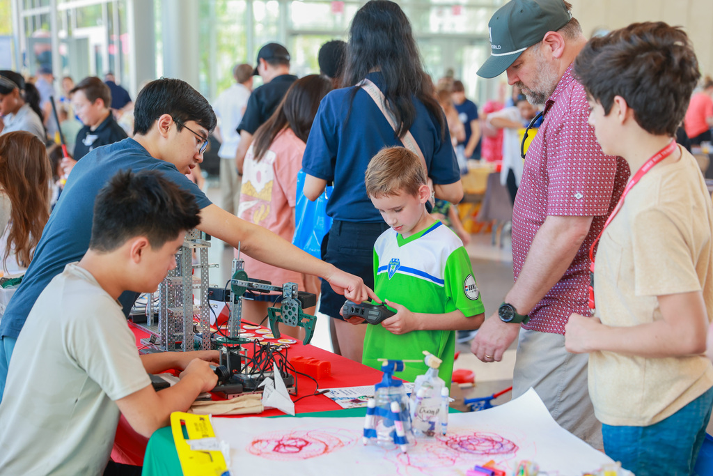 Students and adults gather around a table with Legos and models, examining and interacting with the display.