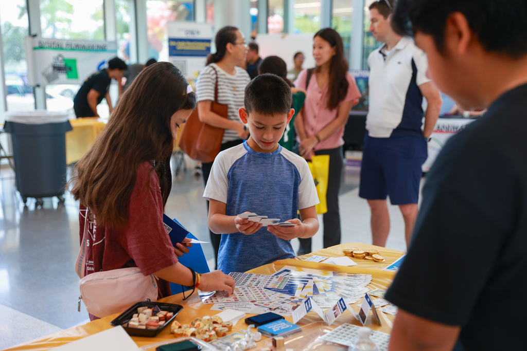 Several students and adults at an event with food and items on a table, inside a building.