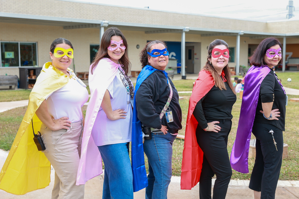 Five women in superhero capes of various colors pose in front of a school building.