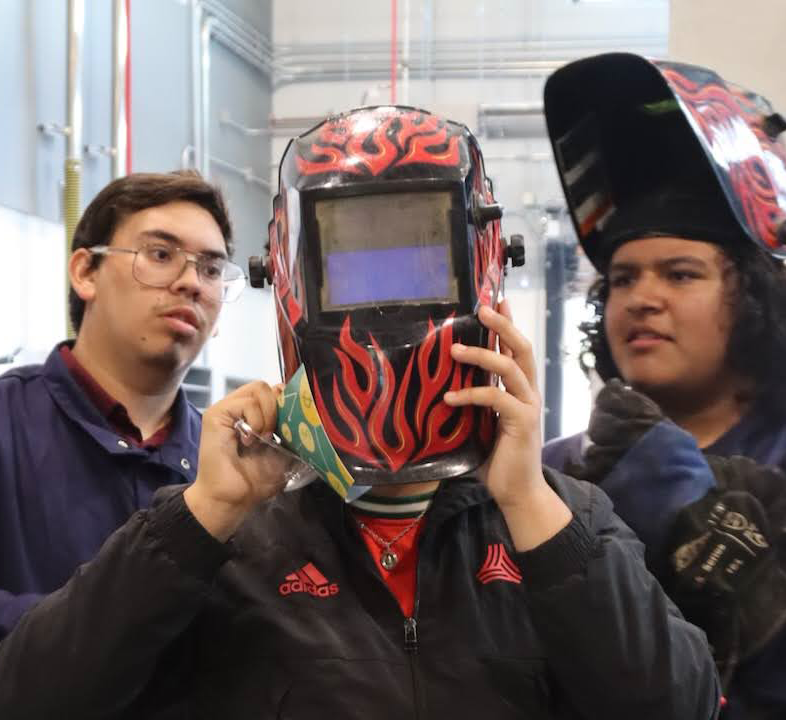 Three young people in a workshop. One is holding a welding mask. Another is wearing a welding mask.