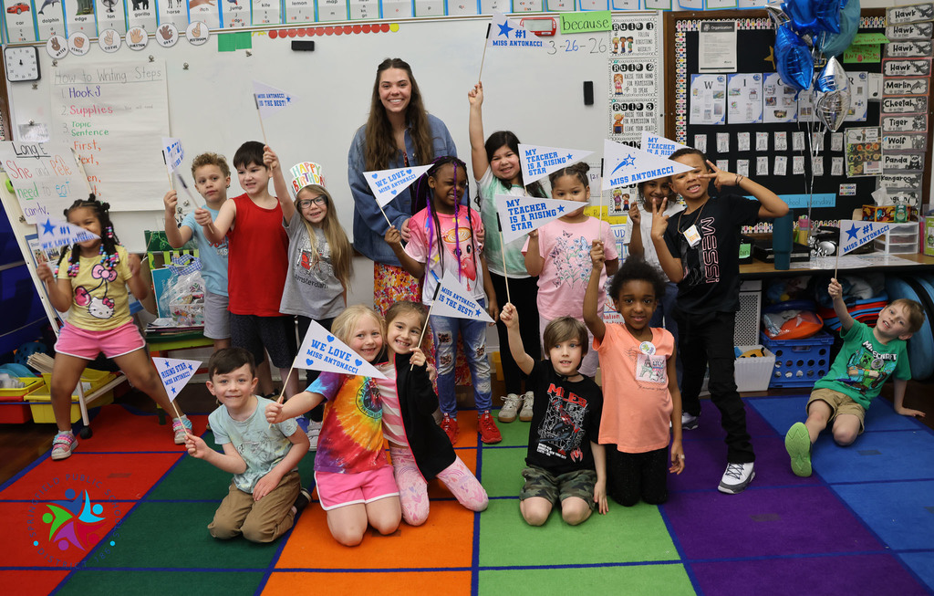 A classroom filled with children holding pennants, a woman stands in the center with a flag, some children crouch on a colorful floor.