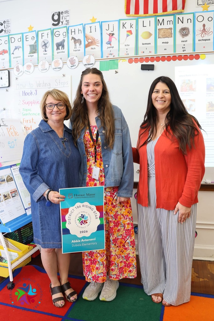 Three women stand in a classroom, one holding a certificate. Behind them, a wall displays an alphabet chart and classroom supplies. A colorful rug is on the floor.