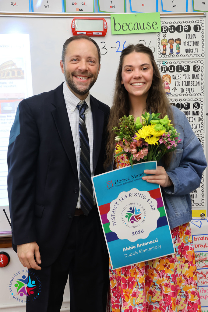 A man and a woman stand together; the woman holds a colorful bouquet and a certificate. They stand against a classroom background.