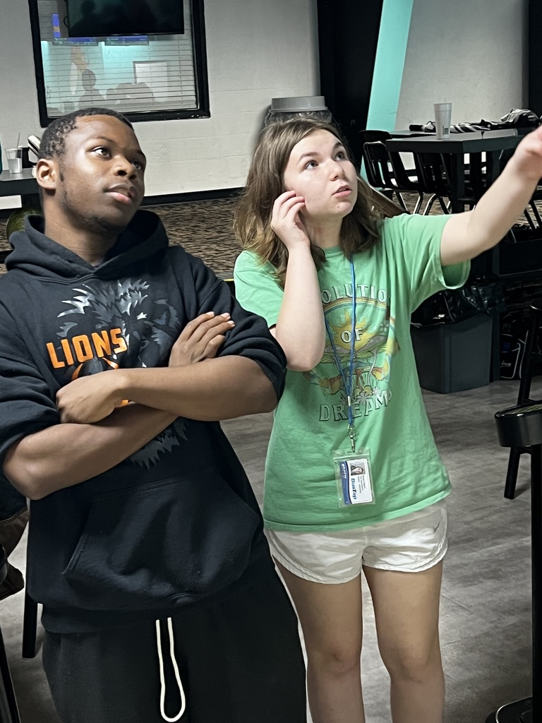 Two people, a man in black and a woman in green, stand in a room with tables and chairs.