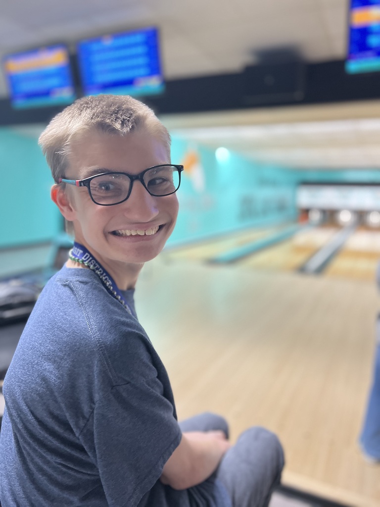 A person in glasses and a blue shirt smiles at the camera in a bowling alley.