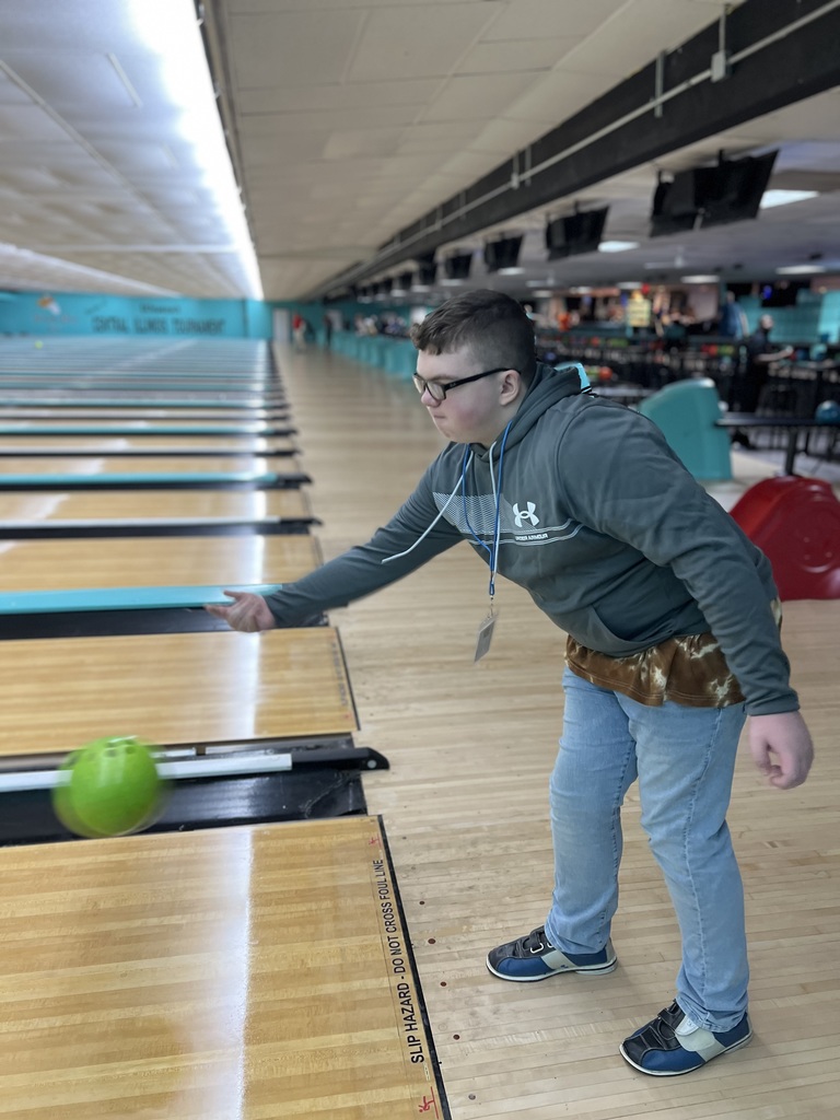 A person in a gray hoodie and blue jeans bowls at a bowling alley, releasing a green ball.
