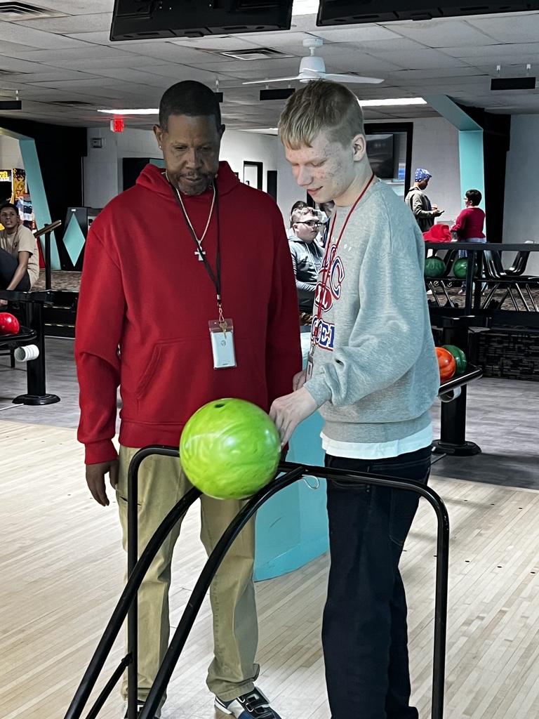 Two men at a bowling alley; one holds a ball, the other stands by a lane.