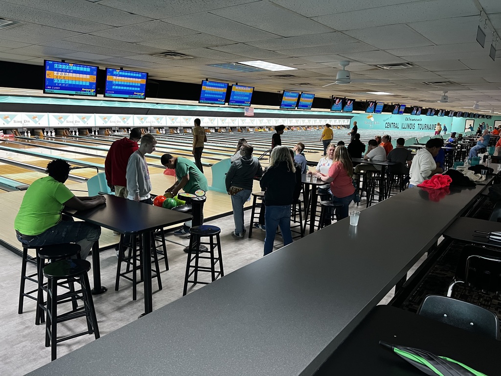 Bowling alley with people sitting at tables. Bowling lanes, scores on screens, and overhead lights.