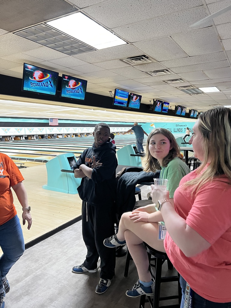 People are in a bowling alley. A person is bowling while two women watch. Monitors are on the wall.