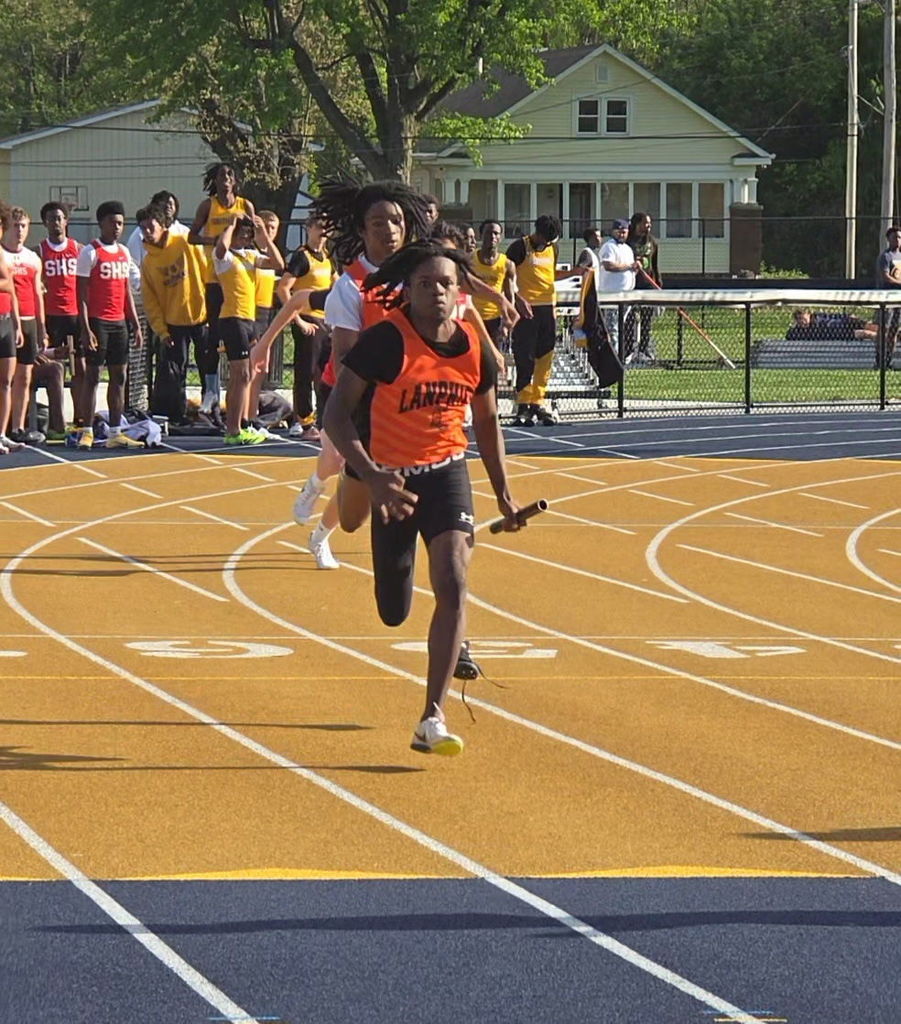 A runner wearing an orange shirt with "LANCE" in white letters sprints on a track. Spectators stand behind the track.