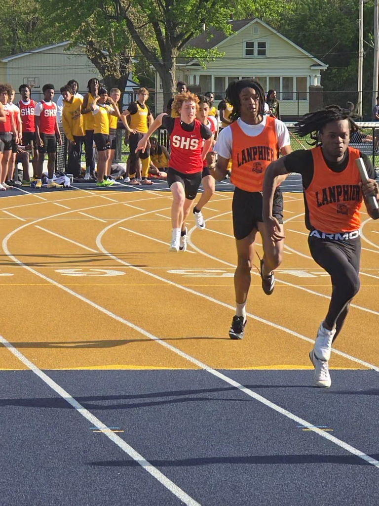 Three athletes sprint on an outdoor track, surrounded by onlookers in the background. Two wear orange vests; one has "LAMPERED" and "ARMO".
