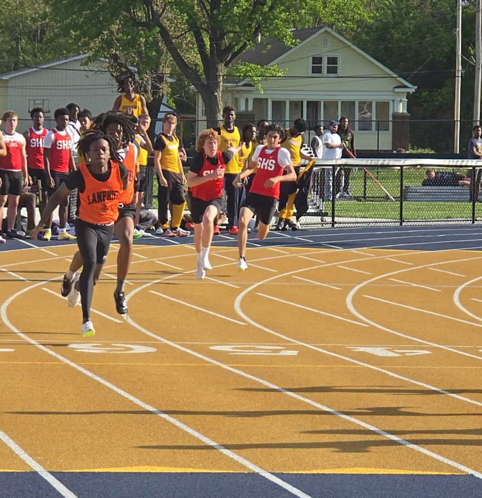Athletes compete on a track field; lanes marked in white and orange. Spectators stand behind a barrier.