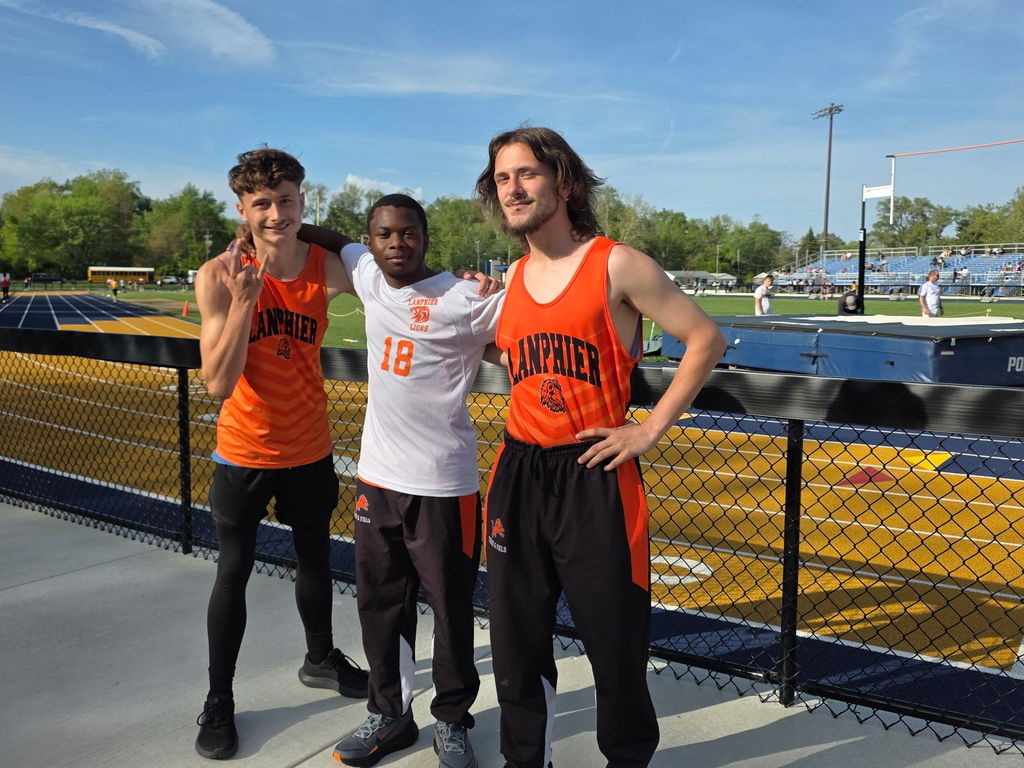 Three people stand on a sidewalk, two in orange tank tops, one in a white shirt. Behind them is a track with a fence.