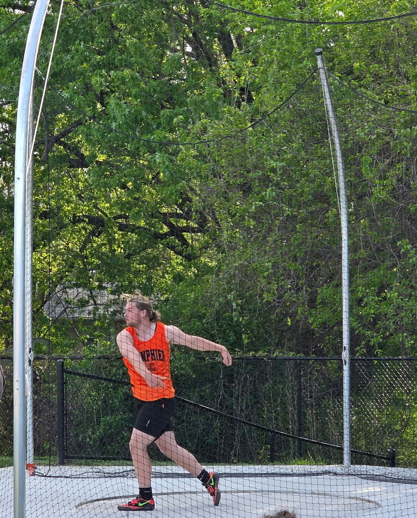 Athlete in orange top and black shorts throws a discus, surrounded by a fence and greenery.