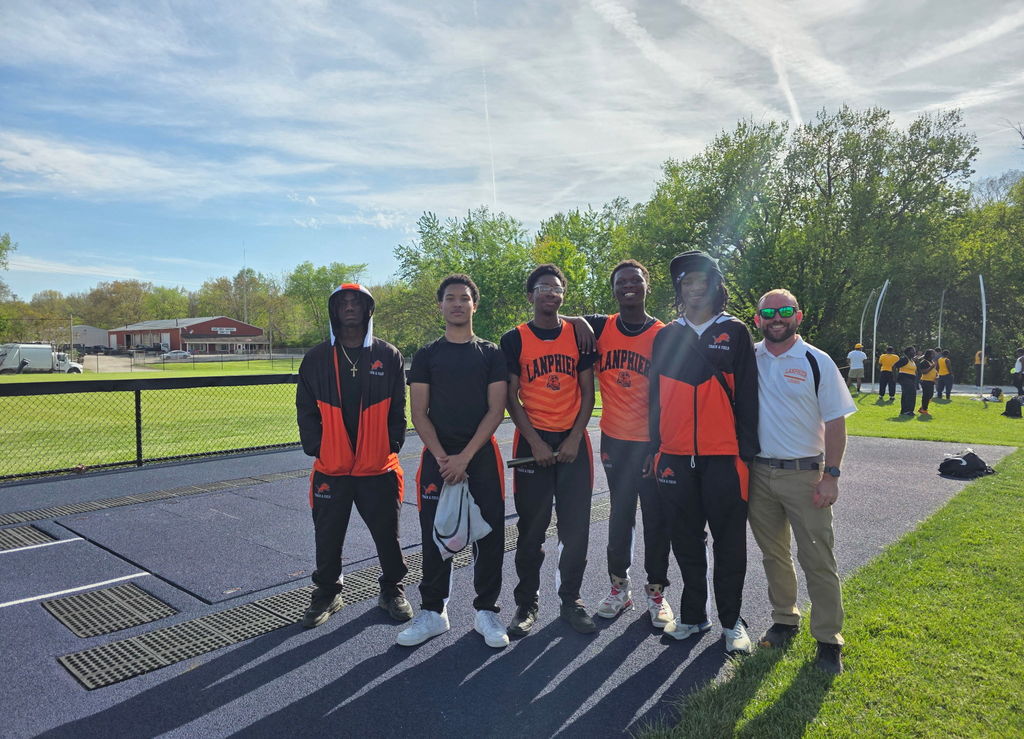 Six people stand in orange and black uniforms on a track with a grassy field in the background.