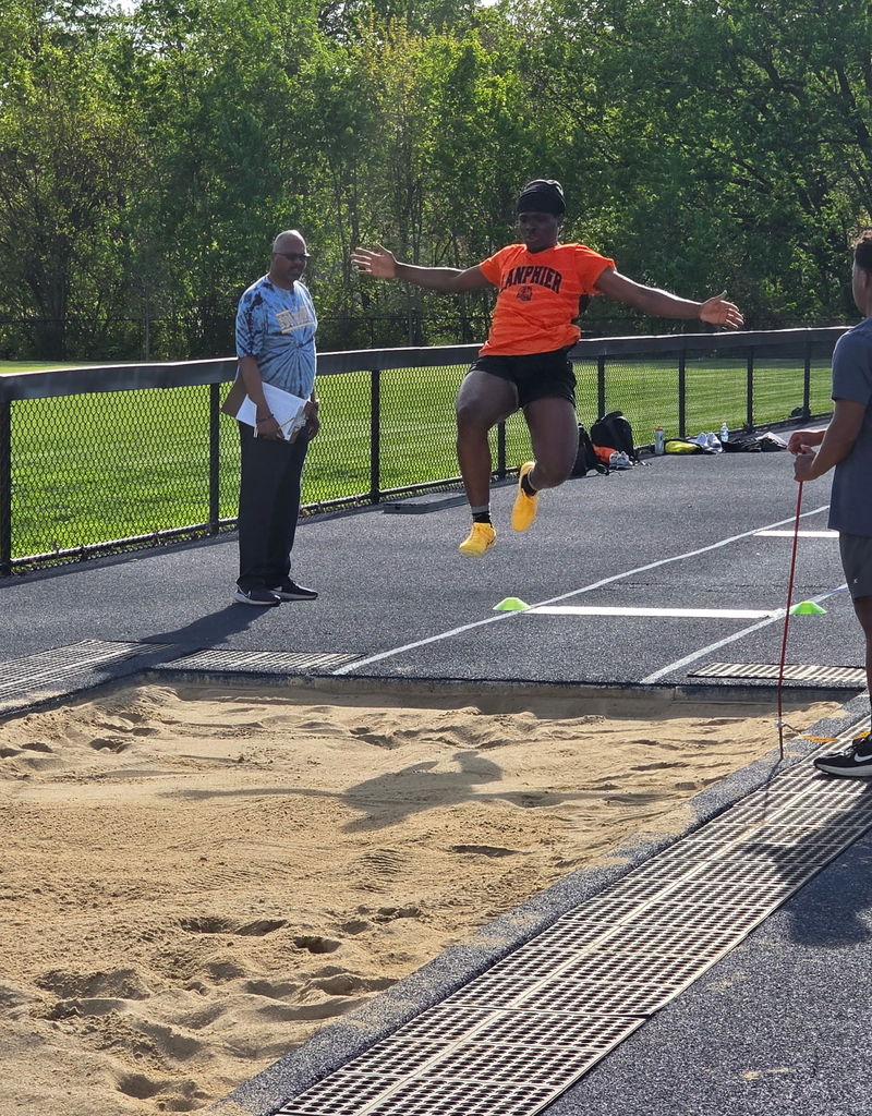Person in orange jumps on a track field, with two people standing behind a fence. Green grass and trees are in the background.