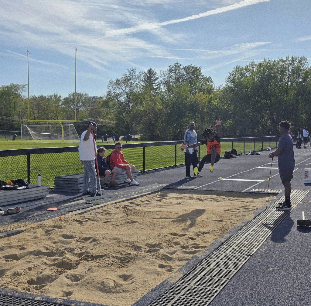 People standing by a long jump sand pit. A person jumps into the pit. The area is surrounded by a fence.