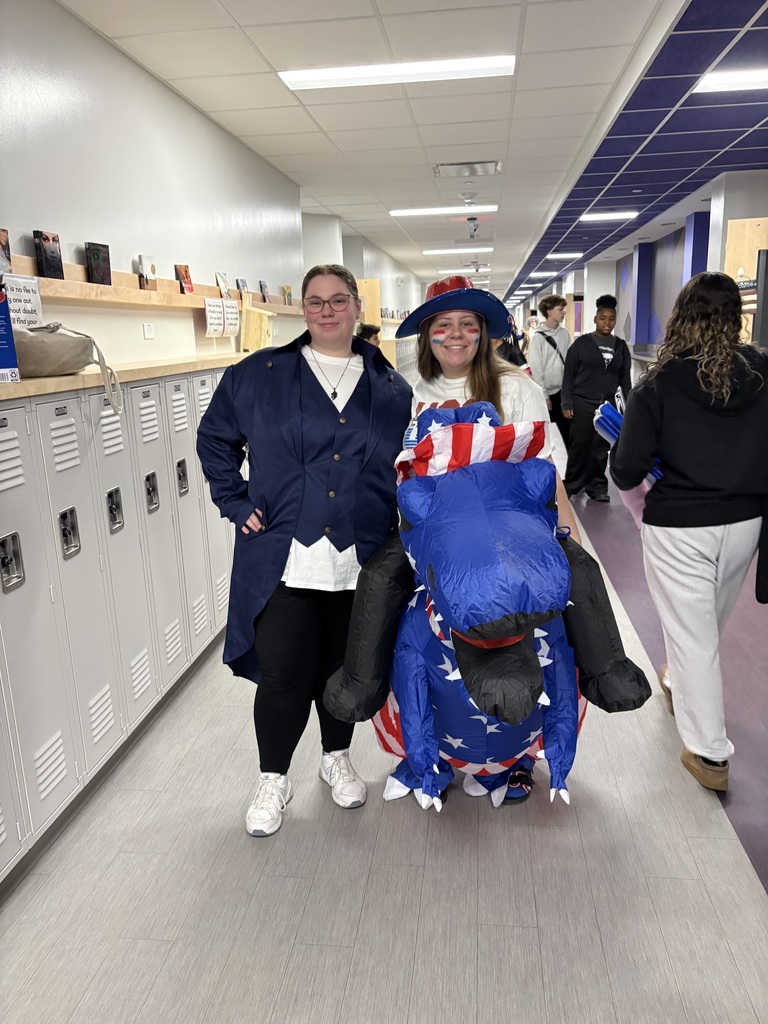 Two people in costumes, one sitting on an inflatable patriotic figure, walk in a hallway lined with lockers.