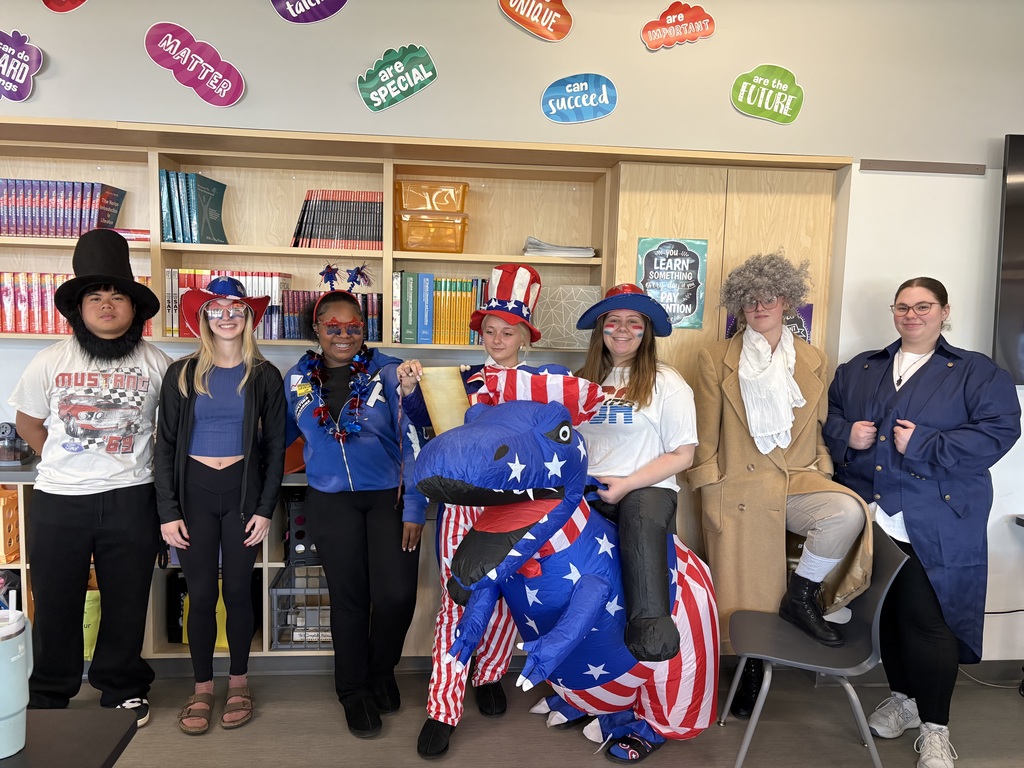 A group of students dressed in costumes posing in a classroom. Colorful decorations adorn the background.