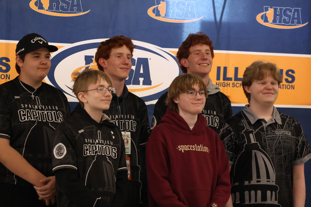 Six boys stand behind a backdrop with logos and text. They wear various colored tops.