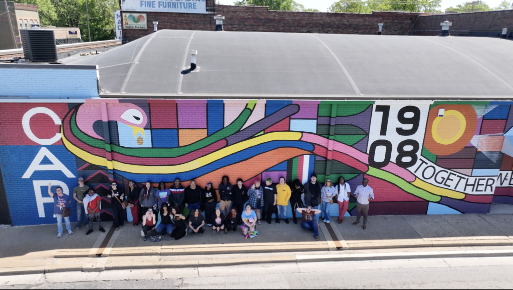 Group of people in front of a colorful mural on a building with text.