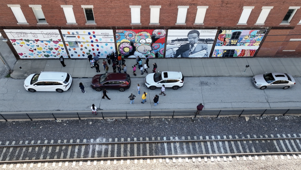 An aerial view of a sidewalk with parked cars, people, and a building with large murals.