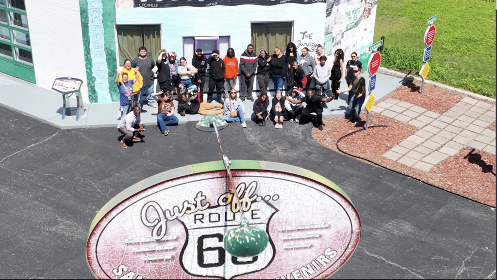 Overhead view of a group standing in front of a building, with a sign reading "Just Old Route 66".