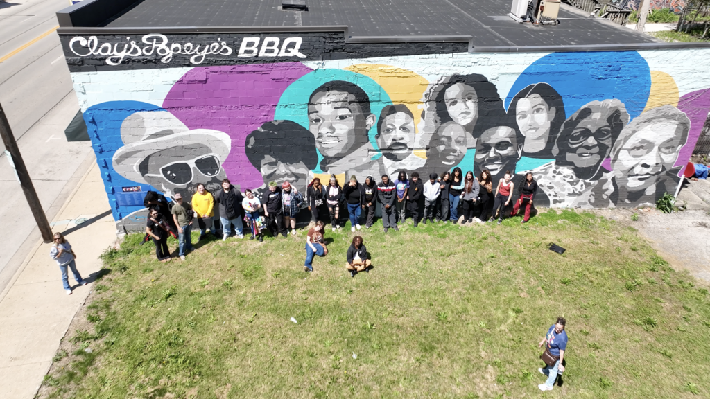 A group of people stand in front of a mural on a building. The mural features several faces.