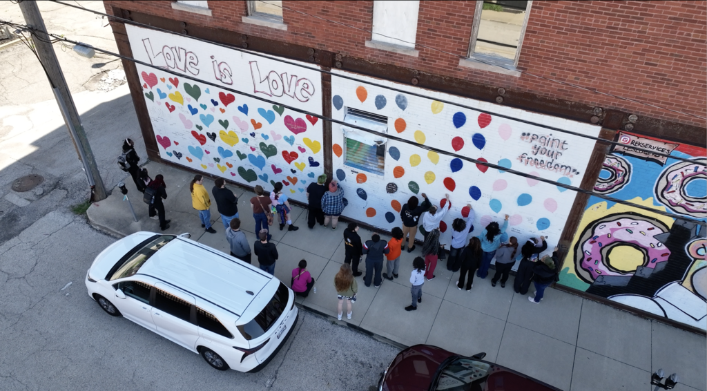 A white SUV parked on the street. People line up at a colorful building mural.