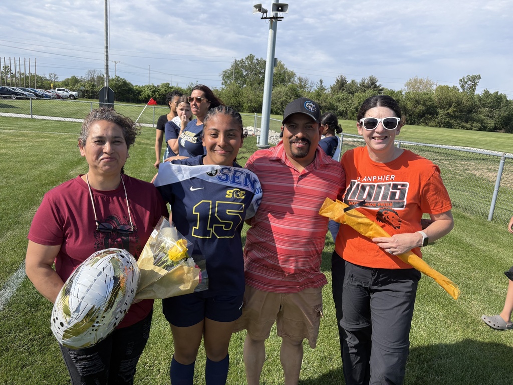 Four adults stand on a grassy field. A girl in a white and blue jersey holds a ball.