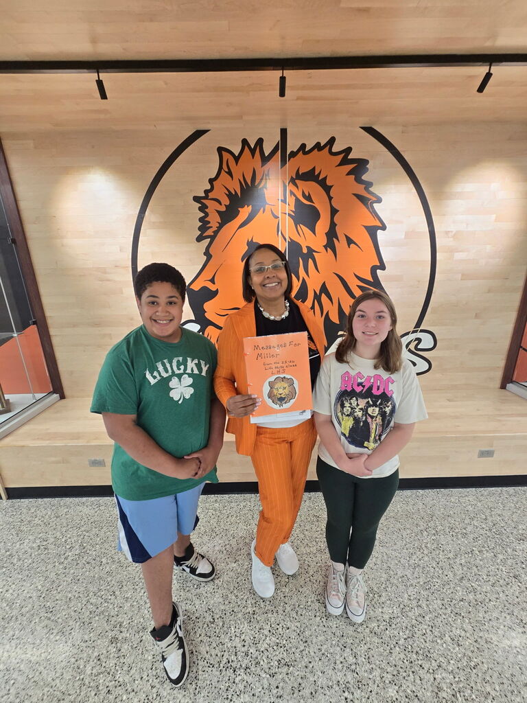 Three students standing in front of a wall with a large lion emblem and a basketball court in the background.