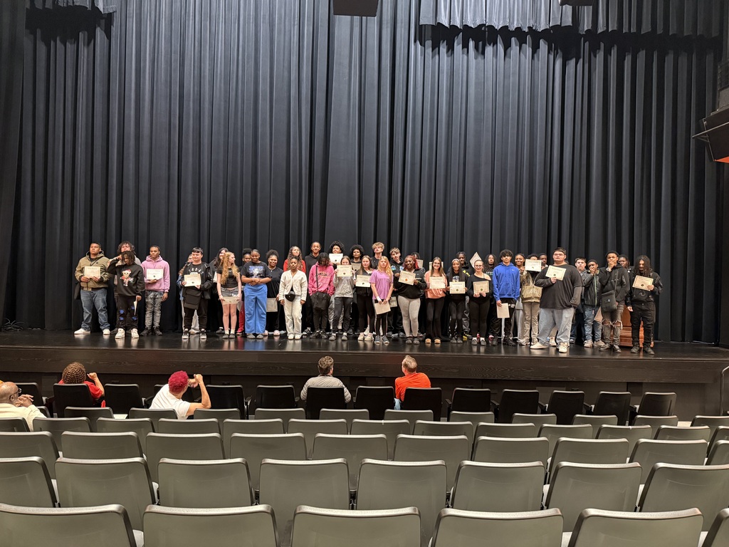 Group of students on stage holding certificates; theater backdrop; rows of empty chairs in foreground.