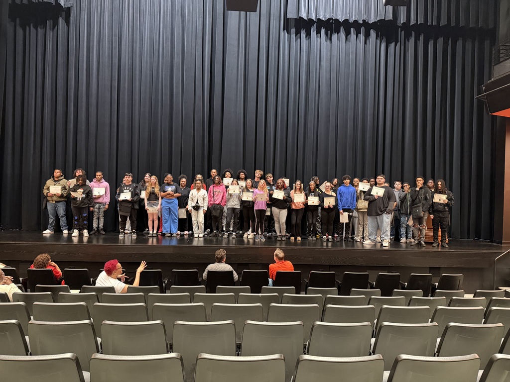 People stand on a stage holding certificates; an audience sits in rows of chairs facing them.
