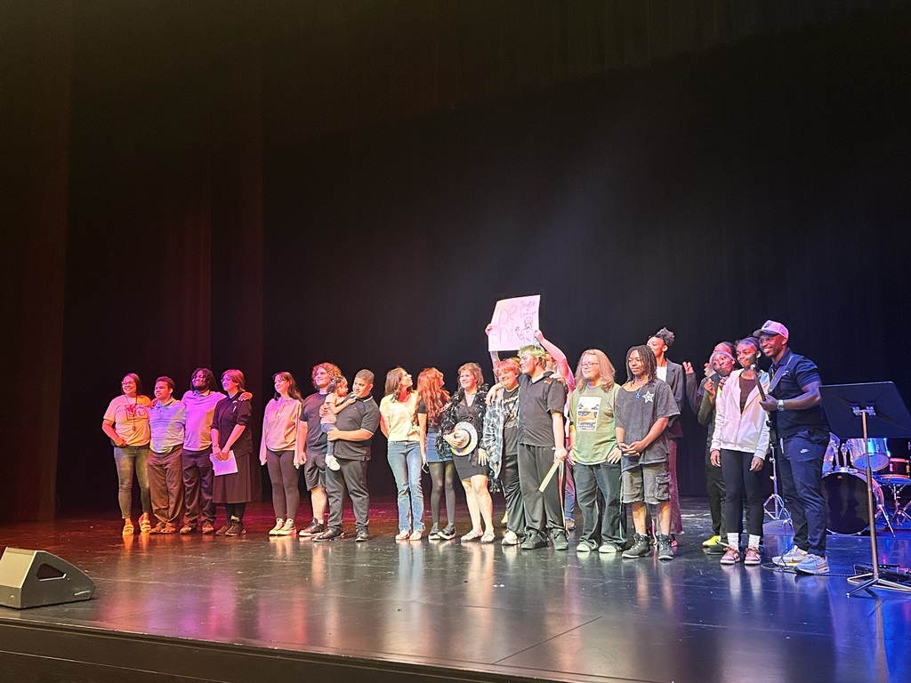 People stand on a stage with one holding a sign. Some wear hats and sneakers. Musical instruments are behind them.