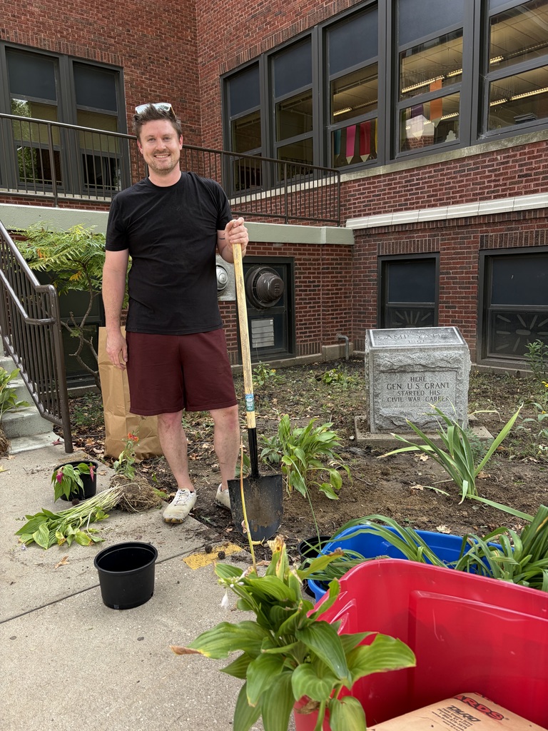 A man holding a shovel stands outside, next to a building, with potted plants and gardening tools.