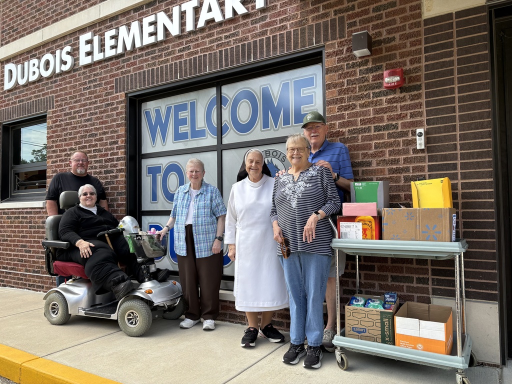 Five people stand outside a building with a sign that reads "Dubois Elementar." A woman in a white dress stands next to a table with boxes.
