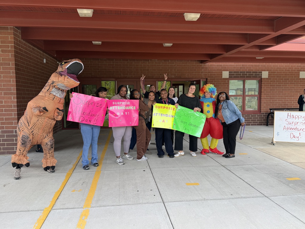 People stand in front of a building holding signs. Some wear costumes. A brick wall and a red roof are visible.
