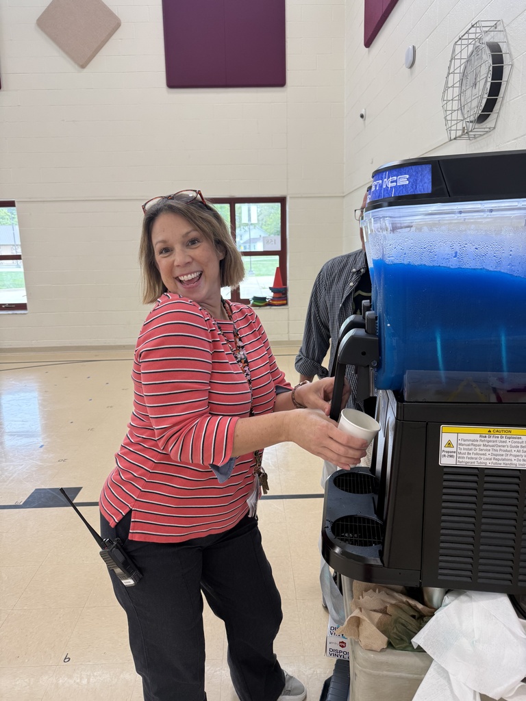 A woman stands next to a beverage machine with a blue container, holding a cup, smiling, and wearing a striped shirt.