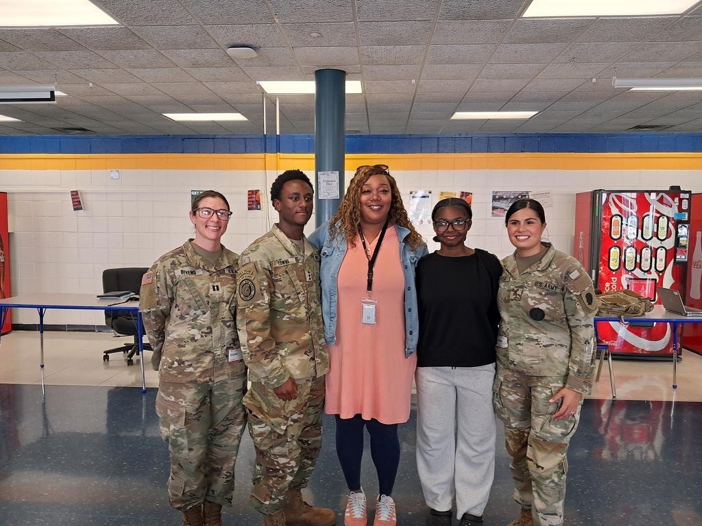 Five adults in uniforms standing in a school hallway. One in a pink dress, three in military uniforms, and one in a black shirt.